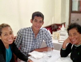 Una mujer, su hijo adulto y su madre se sientan alrededor de una mesa de comedor, todos sonriendo.